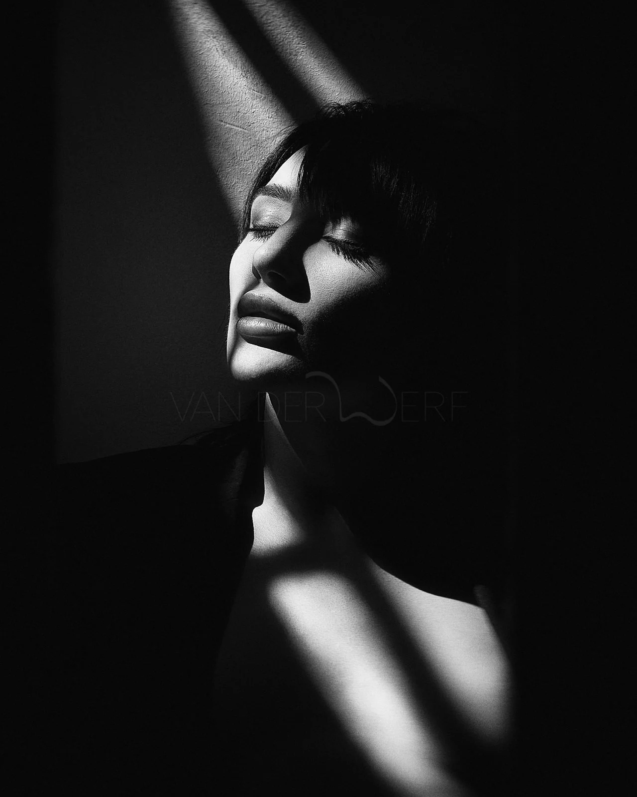 Black and white photo of a woman with eyes closed, smiling slightly, with light and shadows creating diagonal lines across her face and background.