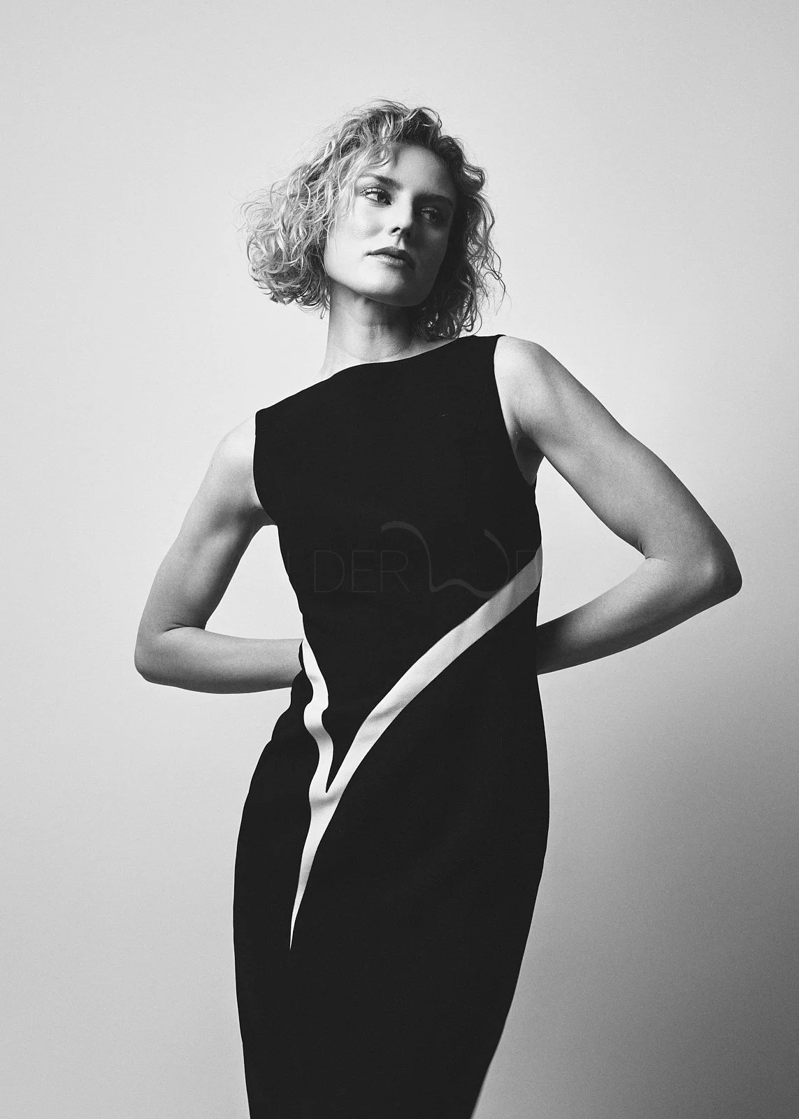 A woman with curly, shoulder-length hair wearing a sleeveless black dress with a white curved stripe design, looking to her left against a plain background.