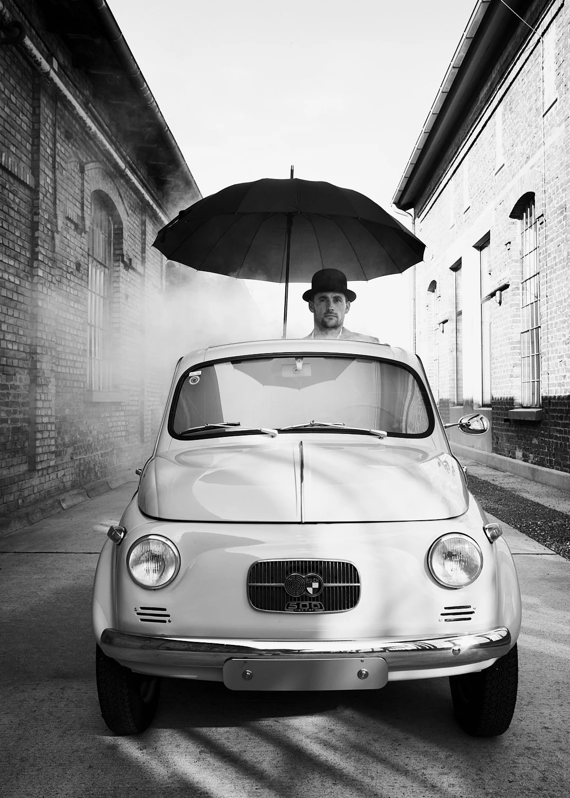 A black-and-white photograph of a vintage small car with a man standing behind it, holding a large umbrella over his head. The man is wearing a bowler hat and looking directly at the camera. The scene is set between brick buildings with barred window