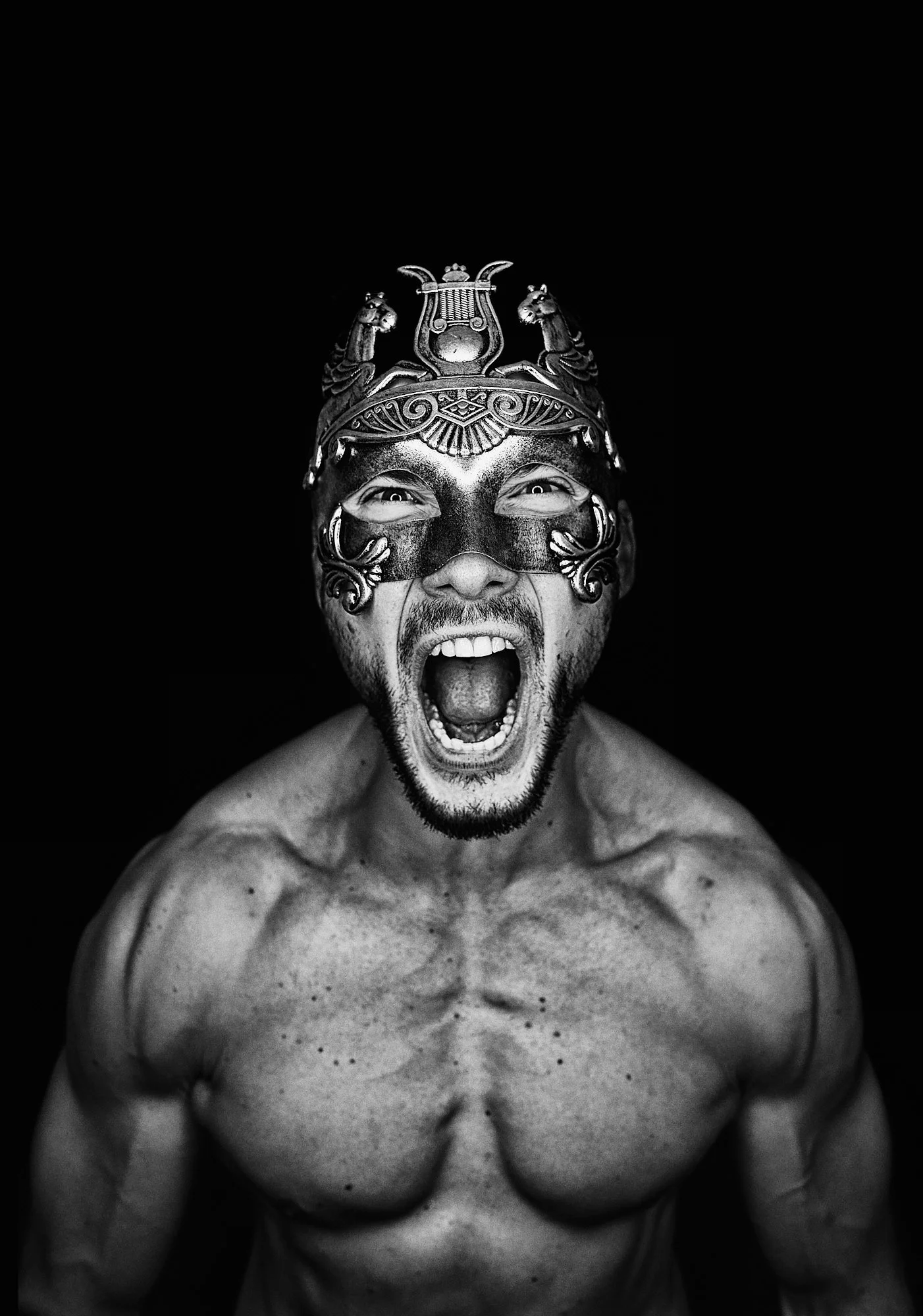 A shirtless man wearing a decorative mask with animal motifs, screaming with mouth open, against a black background.