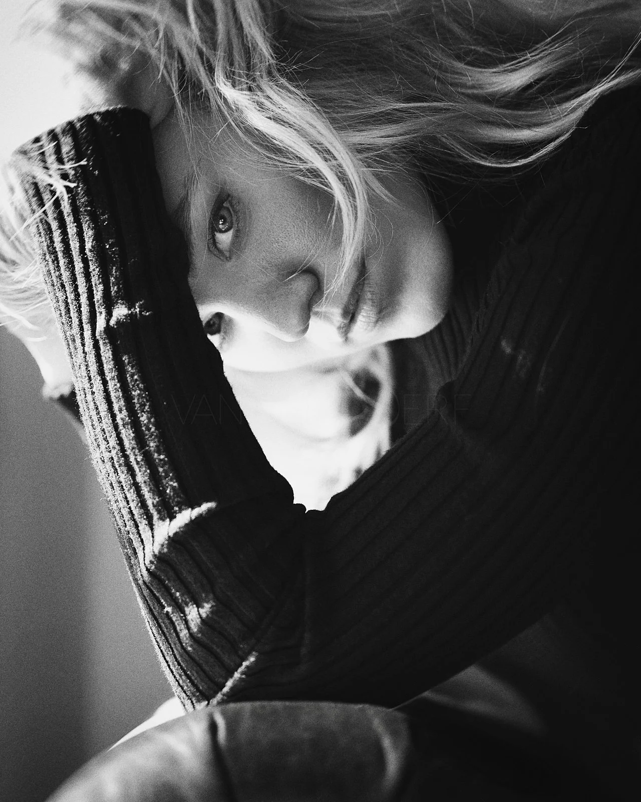 Black and white portrait of a woman with wavy hair partially covering her face, wearing a black hat and a ribbed black top, posed in a close-up shot.