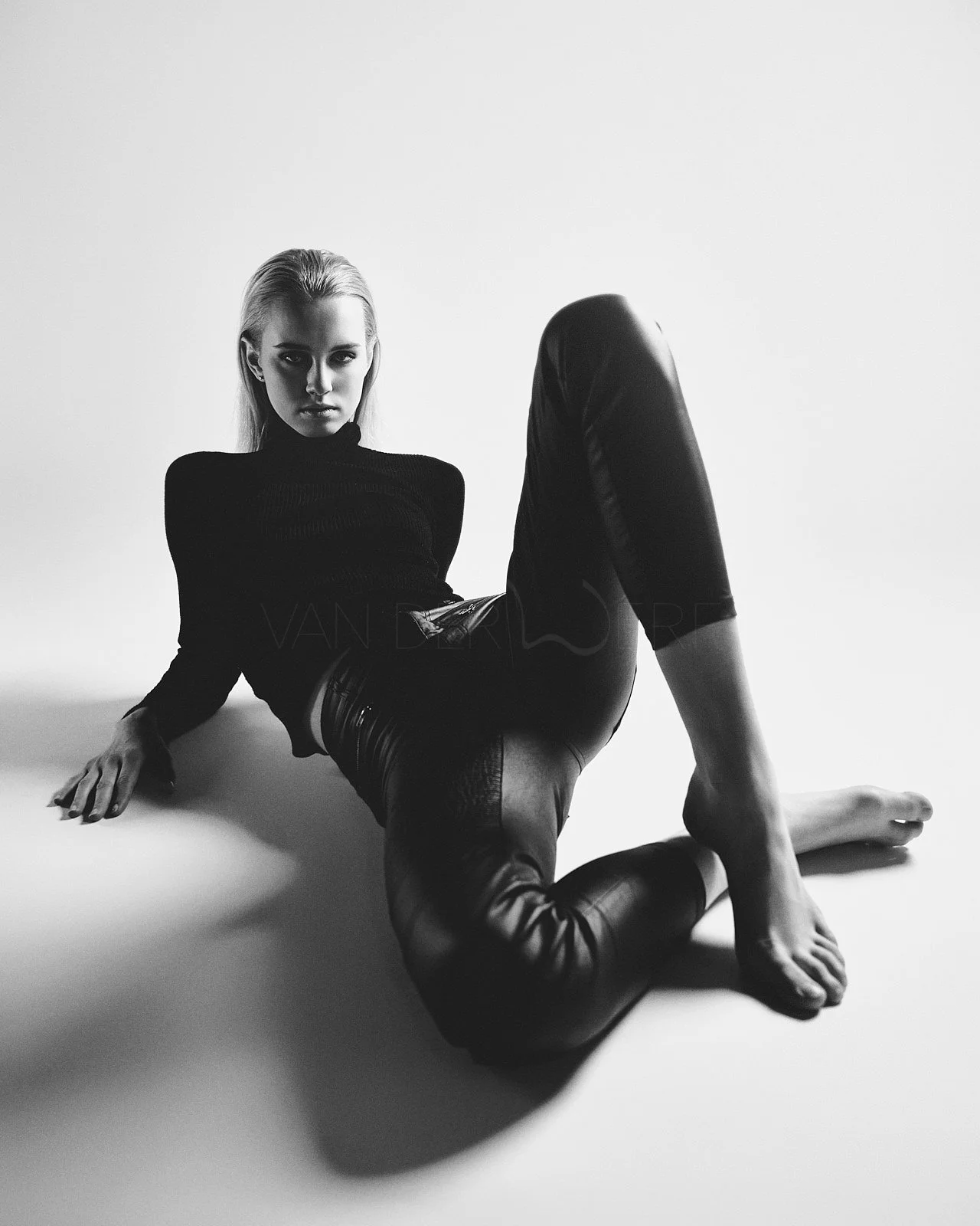 Black and white photo of a woman with platinum blonde hair sitting on the floor with one leg bent and the other extended, wearing a black long-sleeve top and shiny black pants, looking directly at the camera against a plain background.