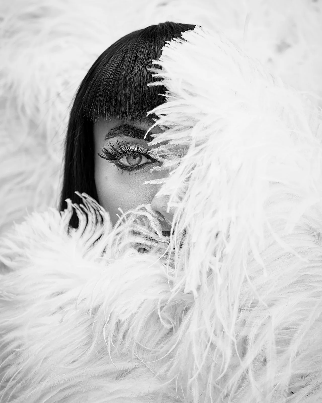 A woman with dark hair and blue eye peeking through white feathers.