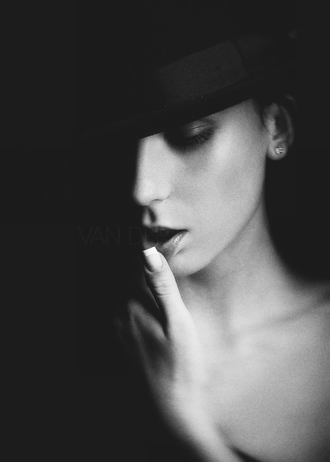 A black and white close-up portrait of a woman wearing a fedora hat, with her eyes closed, finger near her lips, and a single earring visible.