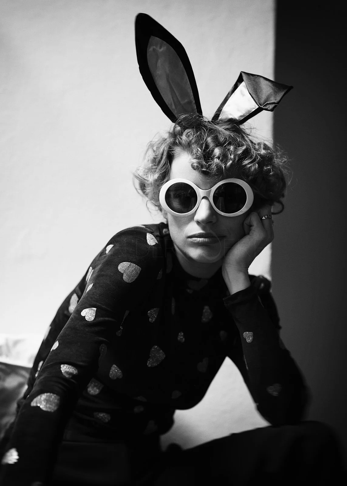A person with curly hair wearing large round sunglasses, bunny ears headband, and a shirt with heart patterns, sitting against a plain wall.