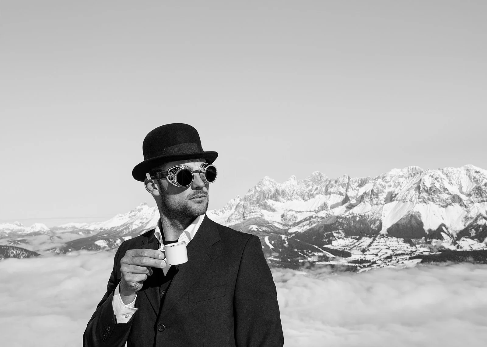 A man in vintage attire, wearing eye goggles, a bowler hat, and a suit, holding a cup of coffee, standing against a backdrop of snow-capped mountains and clouds.