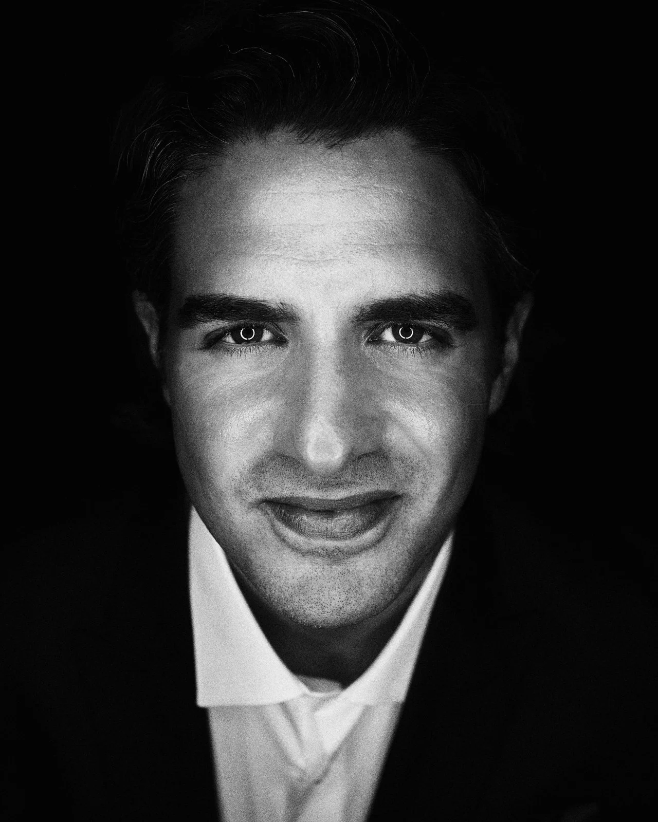Black and white portrait of a man with slicked-back hair, wearing a suit and tie, looking directly at the camera with a slight smile.