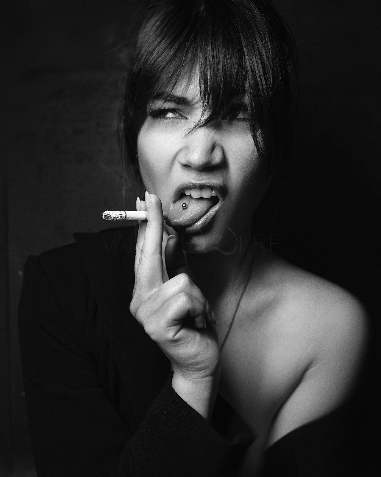 Black and white photo of a woman with short dark hair, making a provocative facial expression, holding a lit cigarette near her mouth with her tongue sticking out, tongue pierced.