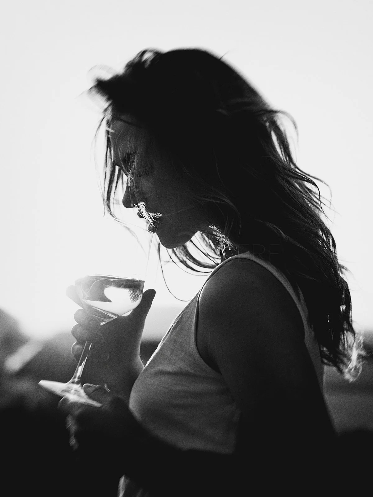 Silhouette of a woman with wavy hair holding a wine glass, backlit with bright light creating a high contrast black and white image.