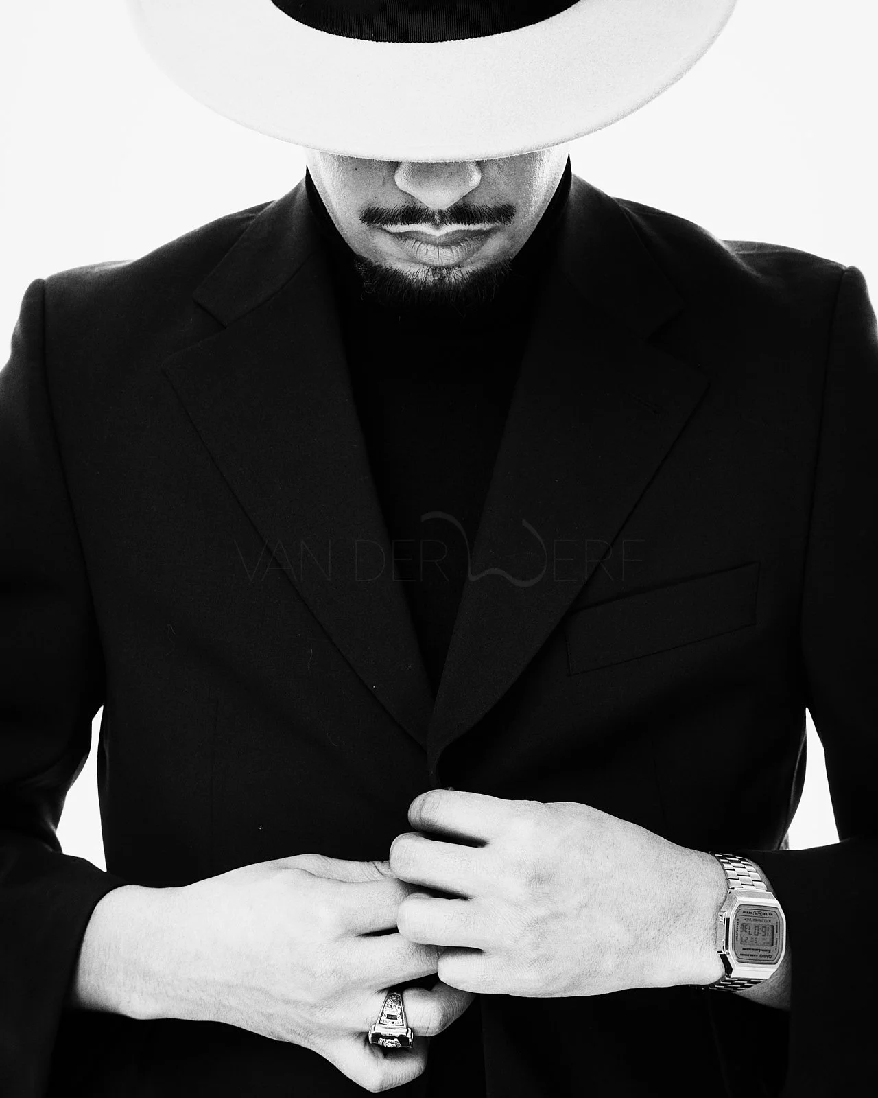 A man in a dark suit and a wide-brimmed hat, with his face partially obscured, is adjusting his suit jacket. He is wearing a watch on his left wrist and a statement ring on his right hand.
