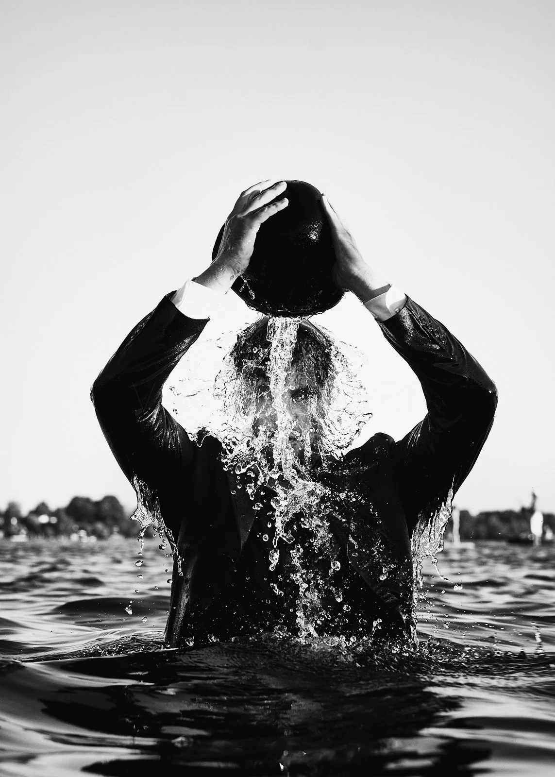 Black and white photo of a person in a suit pouring water over their head in a body of water, with a city skyline in the distance.