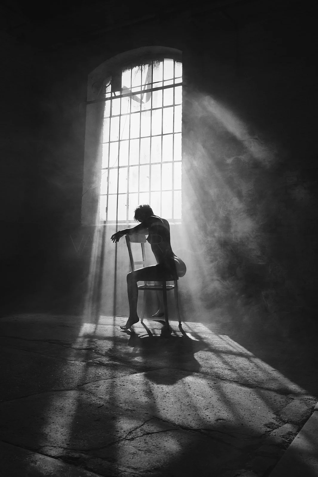 A woman sits on a chair in front of a large window with light streaming in, creating dramatic shadows and misty atmosphere in a dark room.