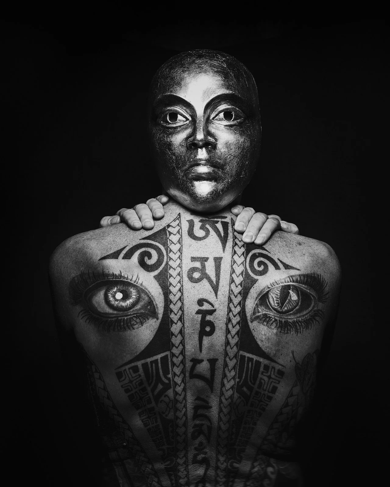 Black and white portrait of a person with an artistic mask covering their face, showing detailed eye features on the mask, with tattoos and symbols on their chest and shoulders, set against a dark background.