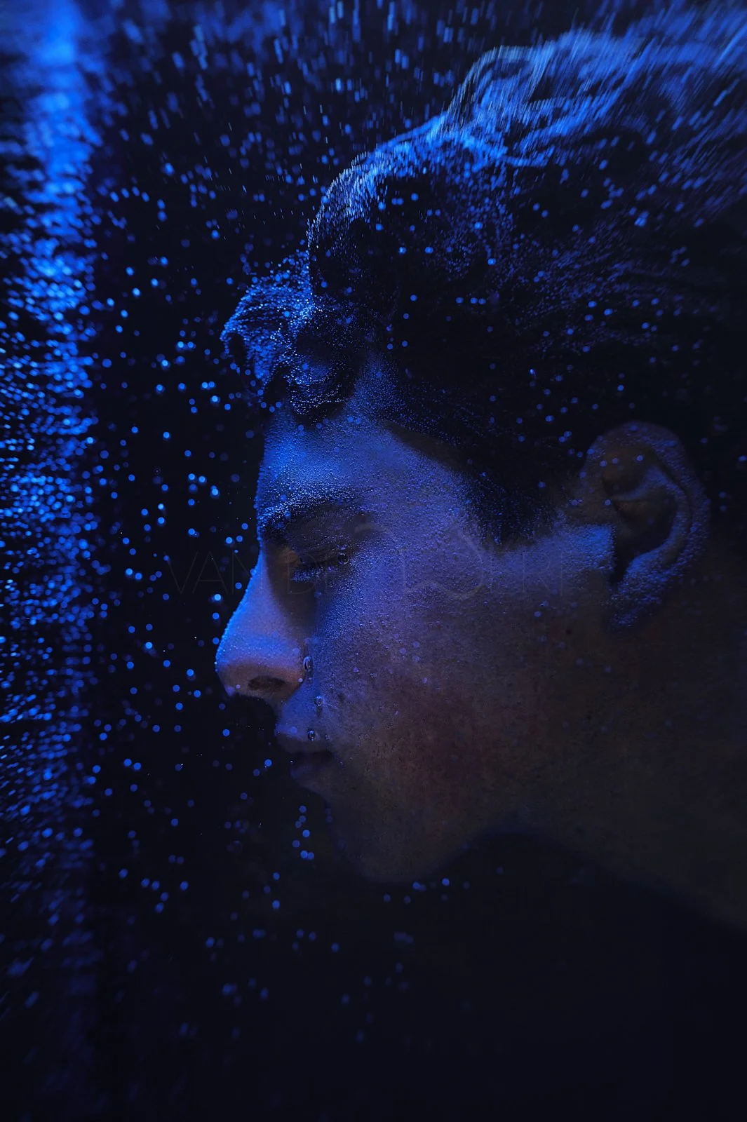 A close-up portrait of a person with closed eyes, surrounded by floating blue particles in a dark environment.
