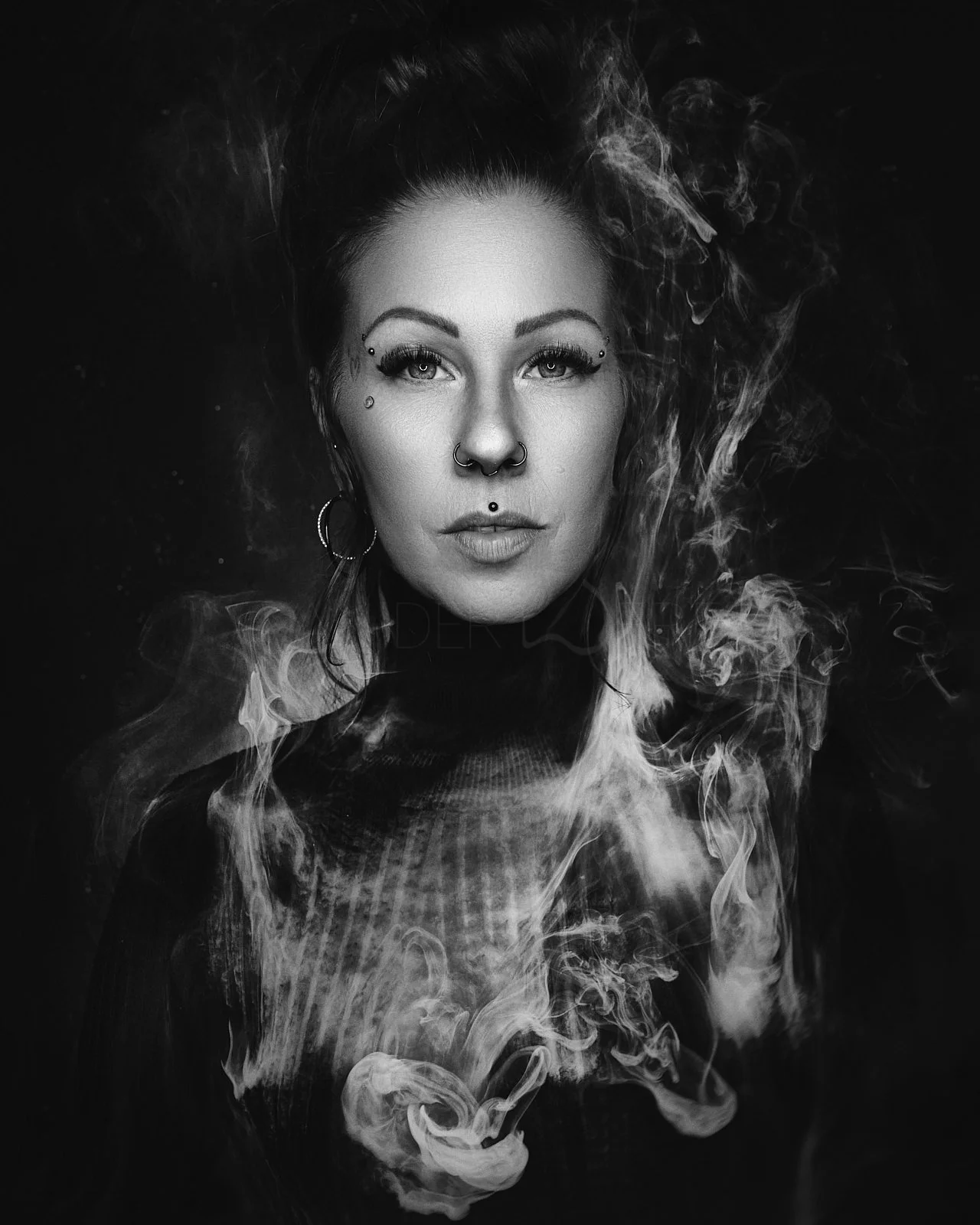 Black and white portrait of a woman with multiple facial piercings, intense eye makeup, and smoke swirling around her, against a dark background.
