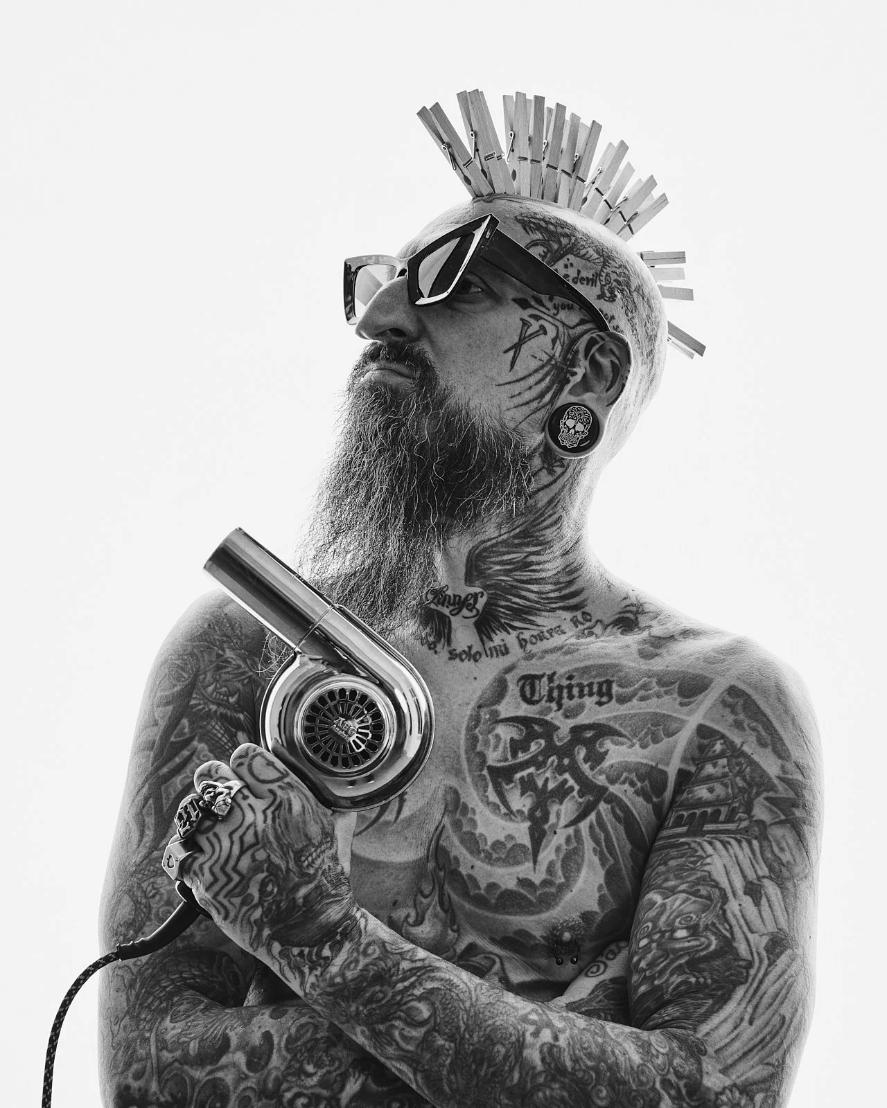 A tattooed man with a beard and glasses, holding a hairdryer to his head, wearing sunglasses, with clothespins on his head, tattoos covering his chest and arms, and large earrings.