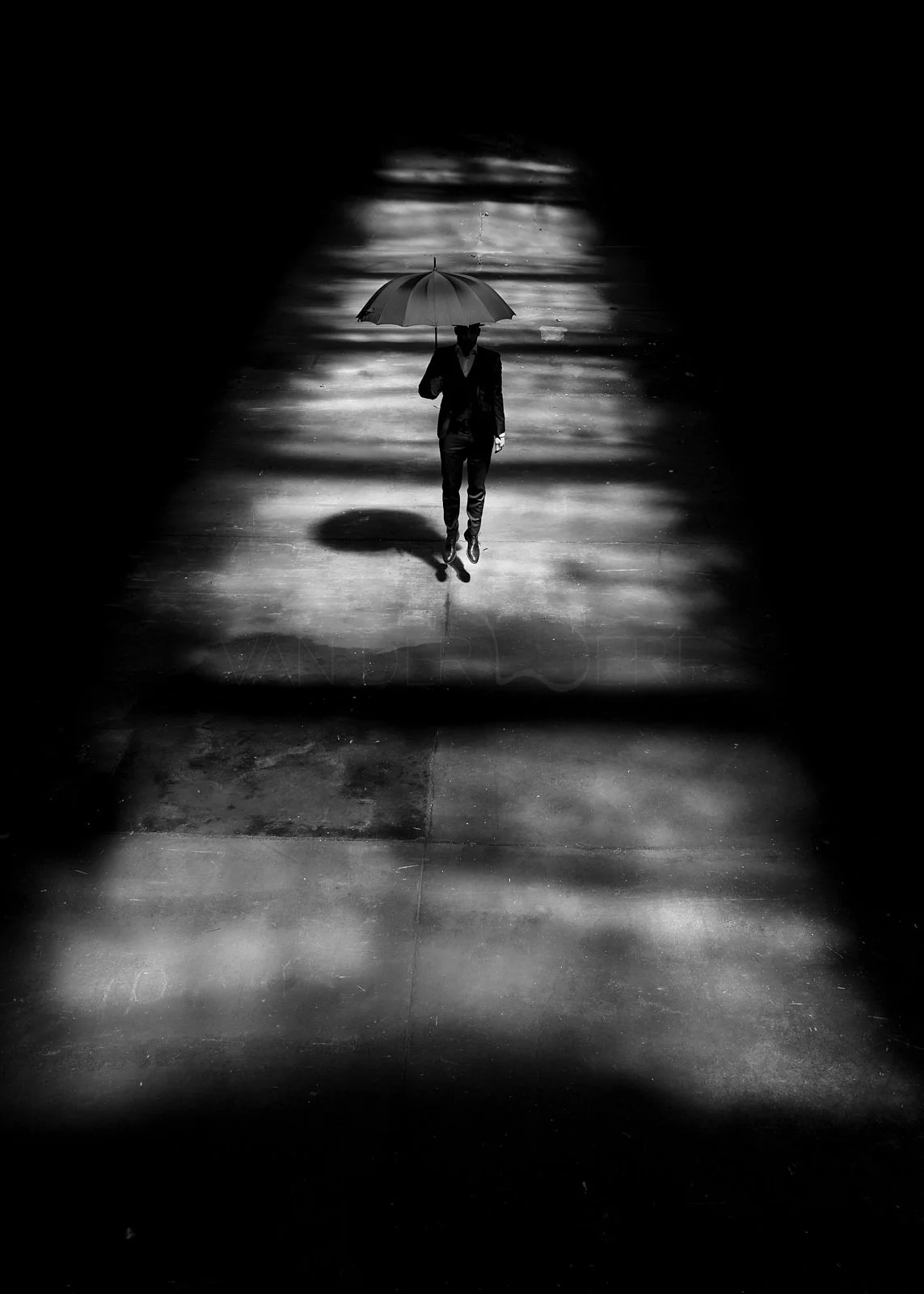 A person in a suit walking on a wet pavement holding an umbrella in a dark, rainy environment, with reflections on the ground and blurred streaks of rain.