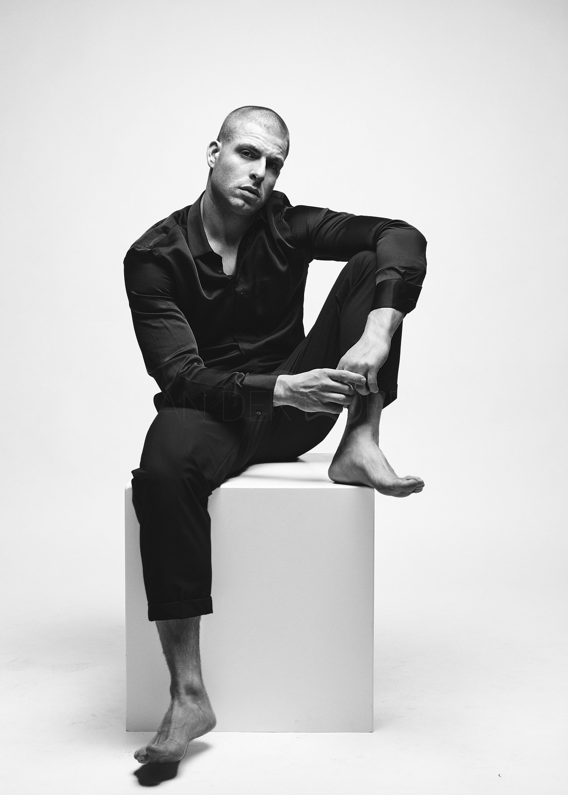 A black and white portrait of a man sitting on a white cube, wearing a black satin shirt and black pants, with one leg crossed over the other and barefoot. He has a shaved head and a serious expression.