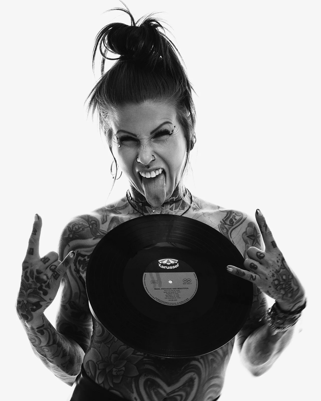 A tattooed woman with her hair in a messy top bun posing with a vinyl record in front of her, sticking her tongue out and making rock hand signs with both hands.