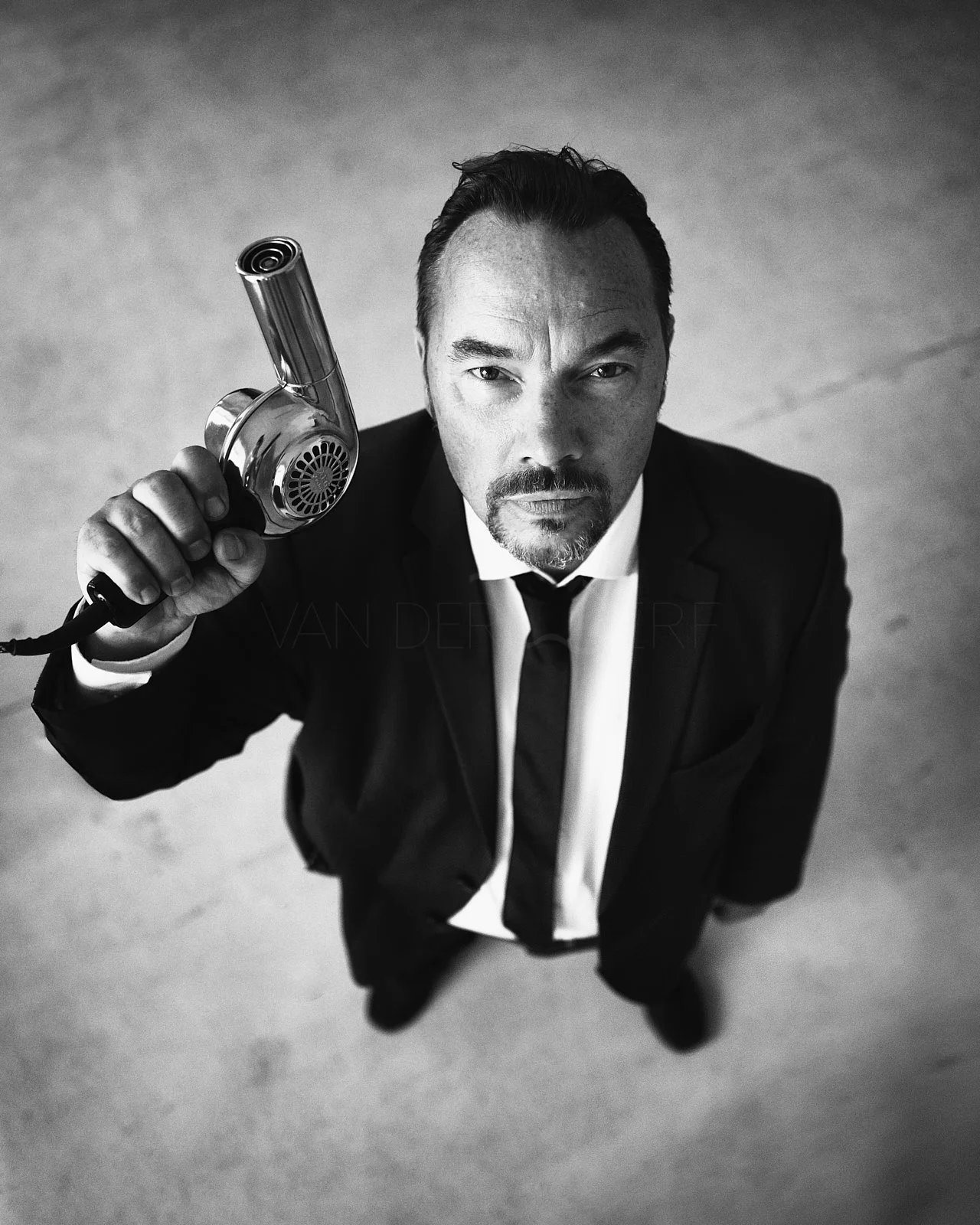 A man in a suit holding a vintage hairdryer pointed at the camera, looking up from a low angle in black and white.