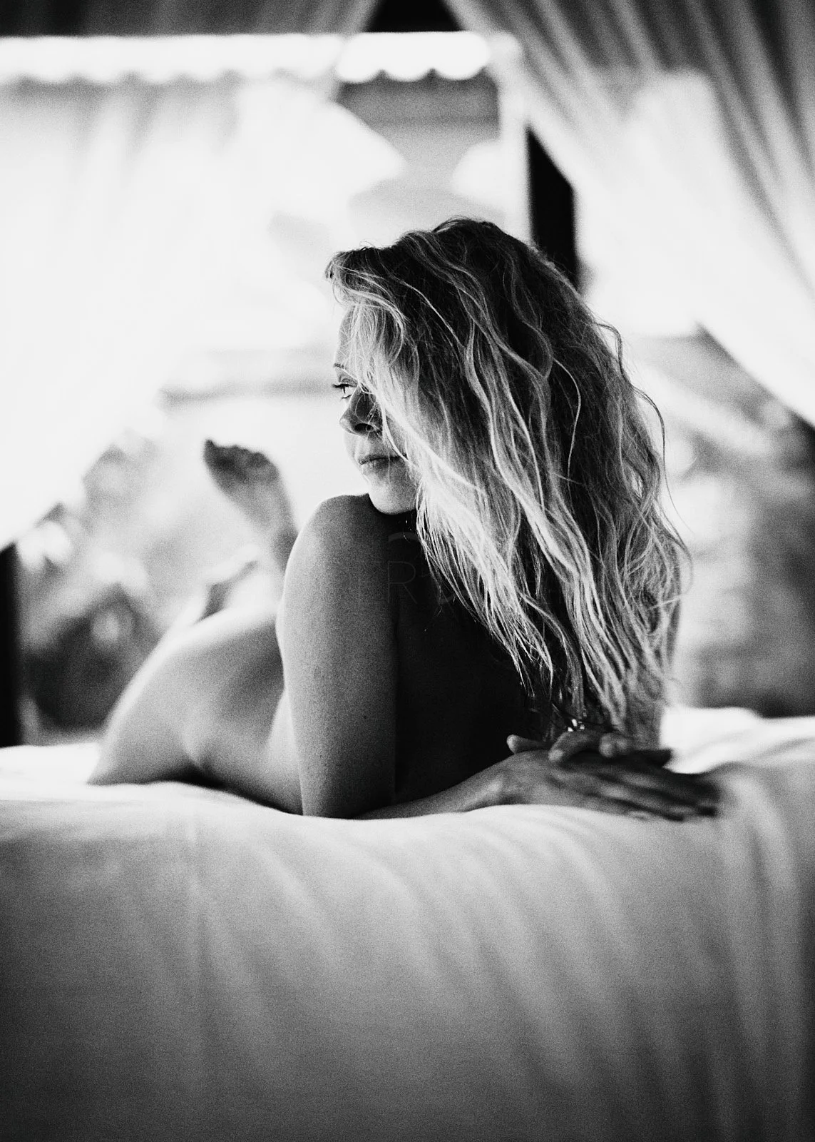 A black and white photo of a woman lying on her stomach on a bed, with long, wavy hair, looking to her left.