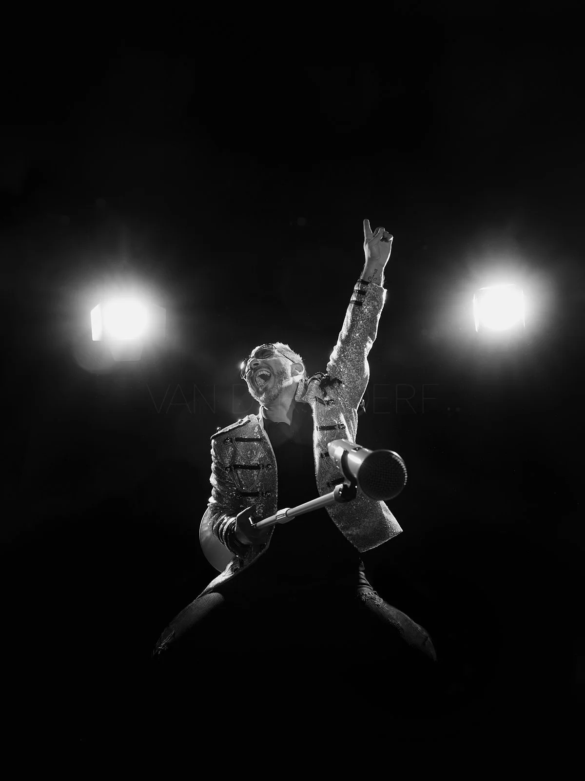 A man playing an electric guitar on stage, smiling and raising one arm with his index finger pointing up, illuminated by bright stage lights, in black and white.