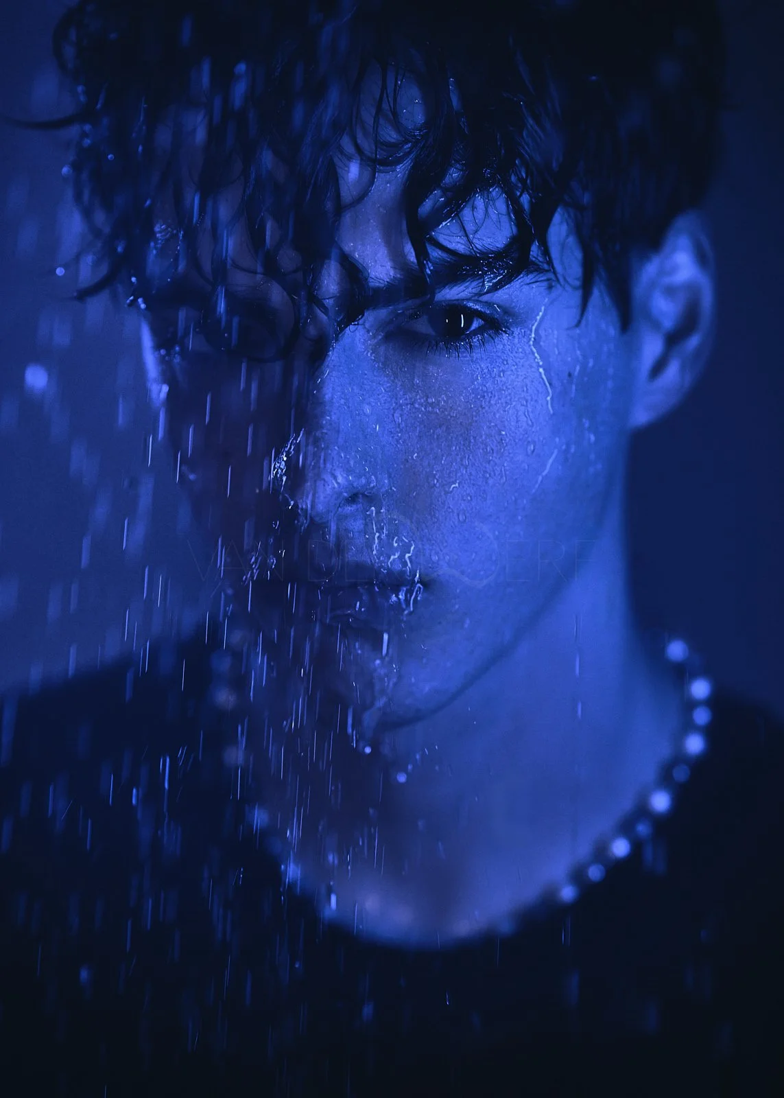 A close-up of a person's face with wet hair and water droplets, illuminated in blue light.
