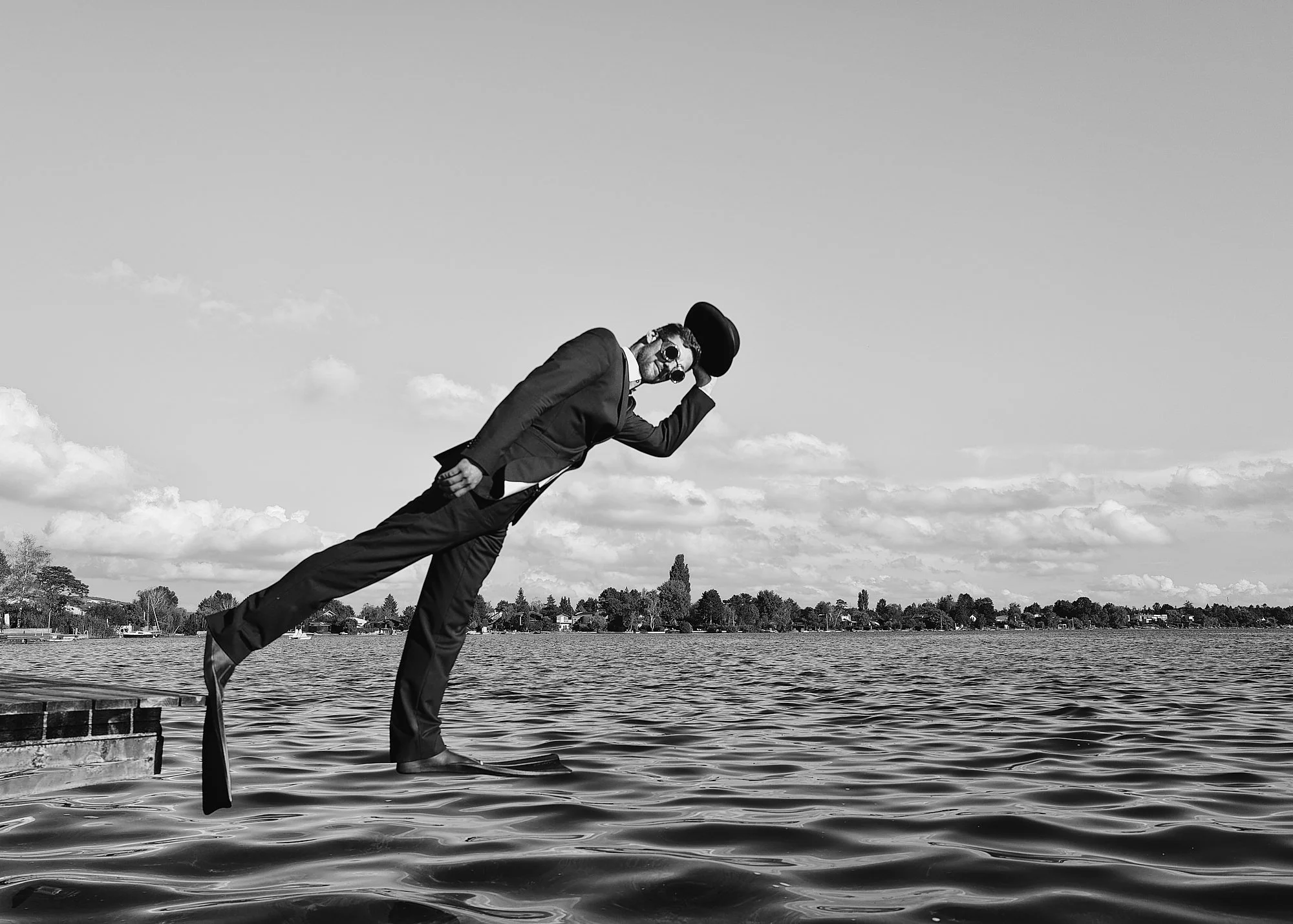 A man in a suit, sunglasses, and a hat, standing on a paddleboard in a body of water, tipping his hat while looking at the camera.