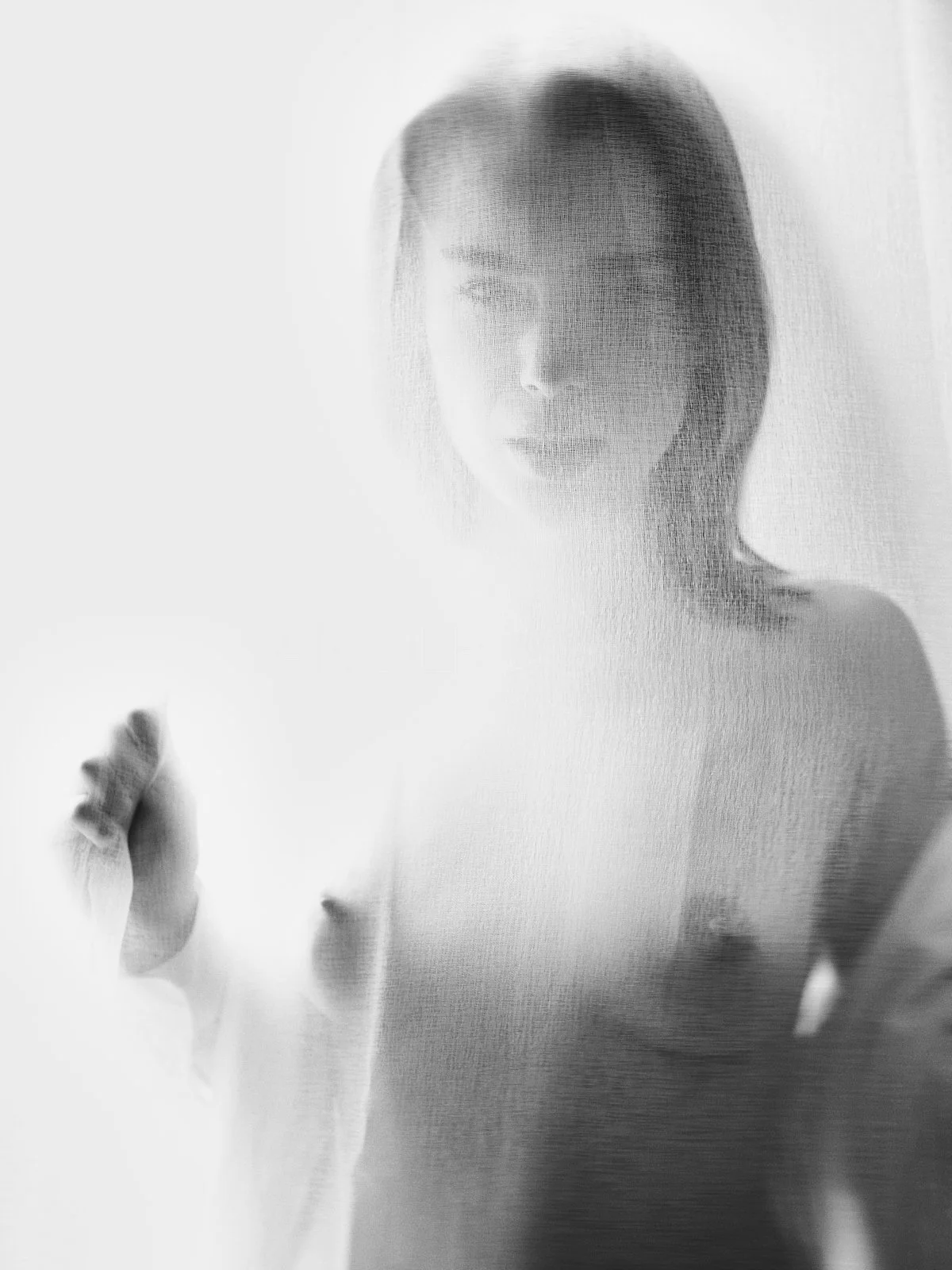A woman with short hair is seen behind a semi-transparent textured curtain or glass, creating a blurred, artistic effect.