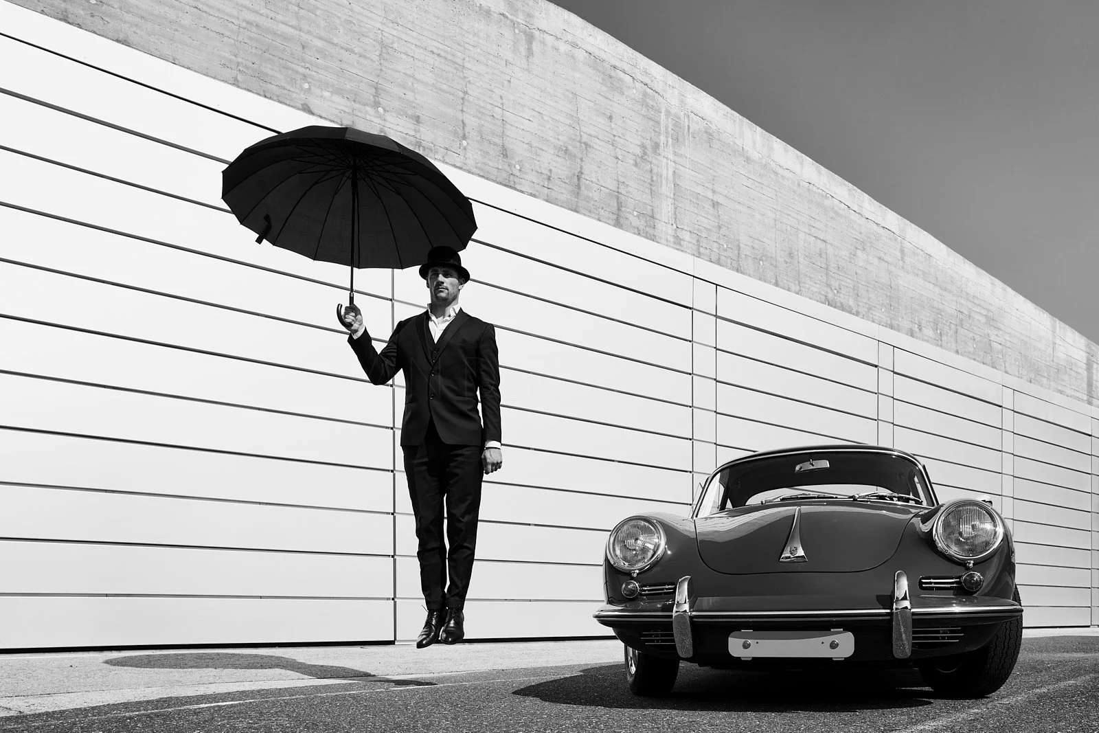 Black and white photo of a man in a suit and bowler hat holding an umbrella, standing next to a vintage sports car in front of a modern building with horizontal siding.