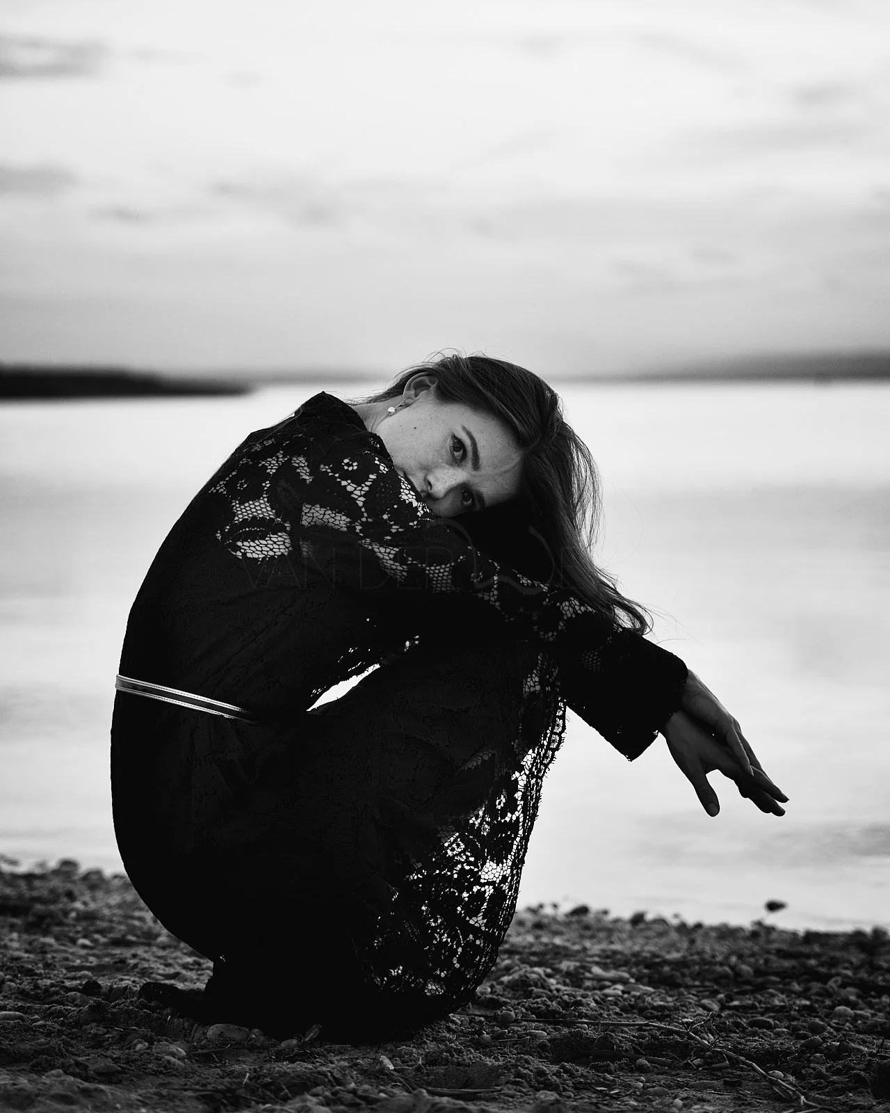 Black and white photo of a woman with long hair, sitting on rocky ground near a body of water, with her knee drawn up and looking at the camera.