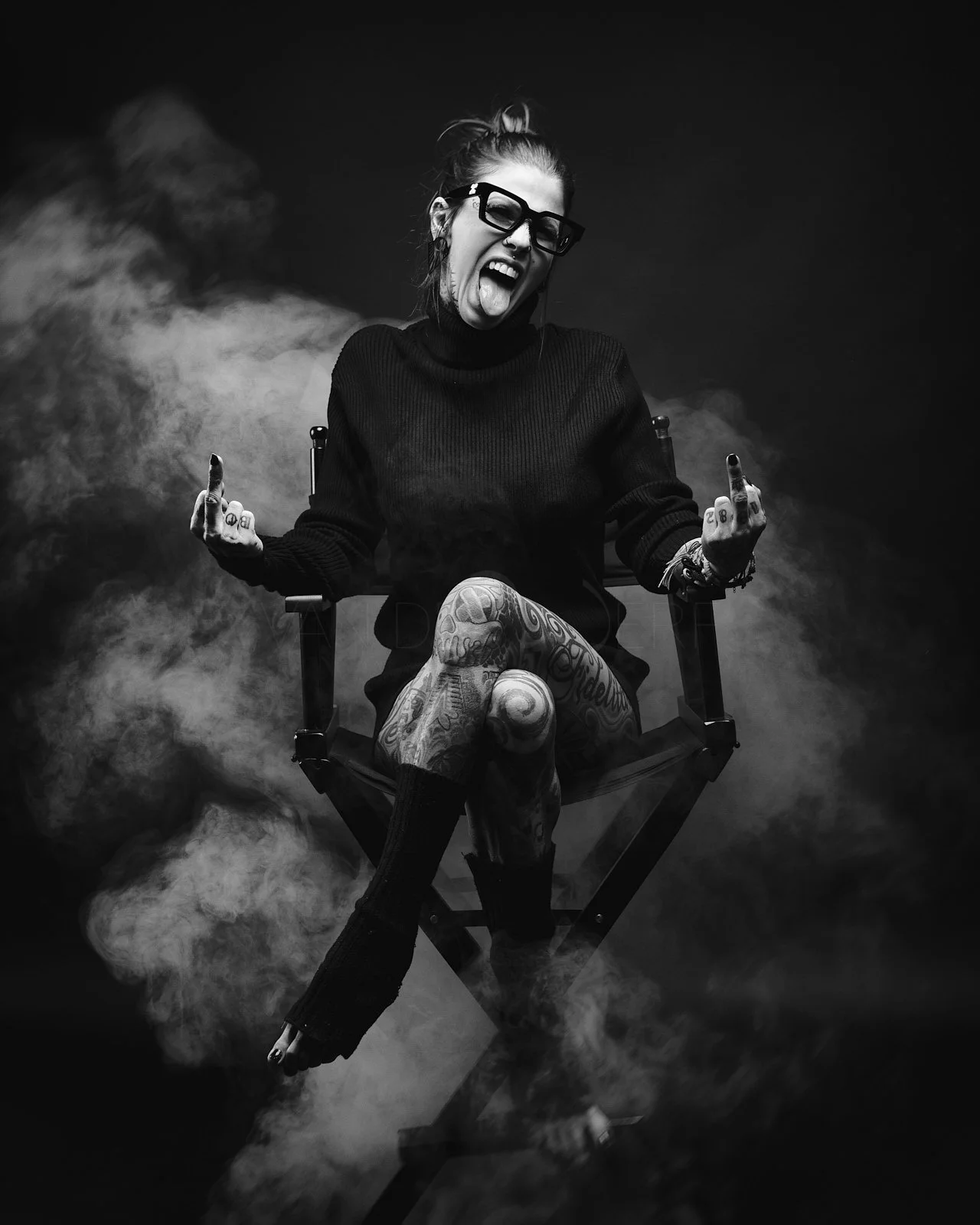 A woman with glasses, tattoos, and her hair in a bun, sitting on a director's chair, sticking out her tongue, with smoke around her in a black-and-white photo.
