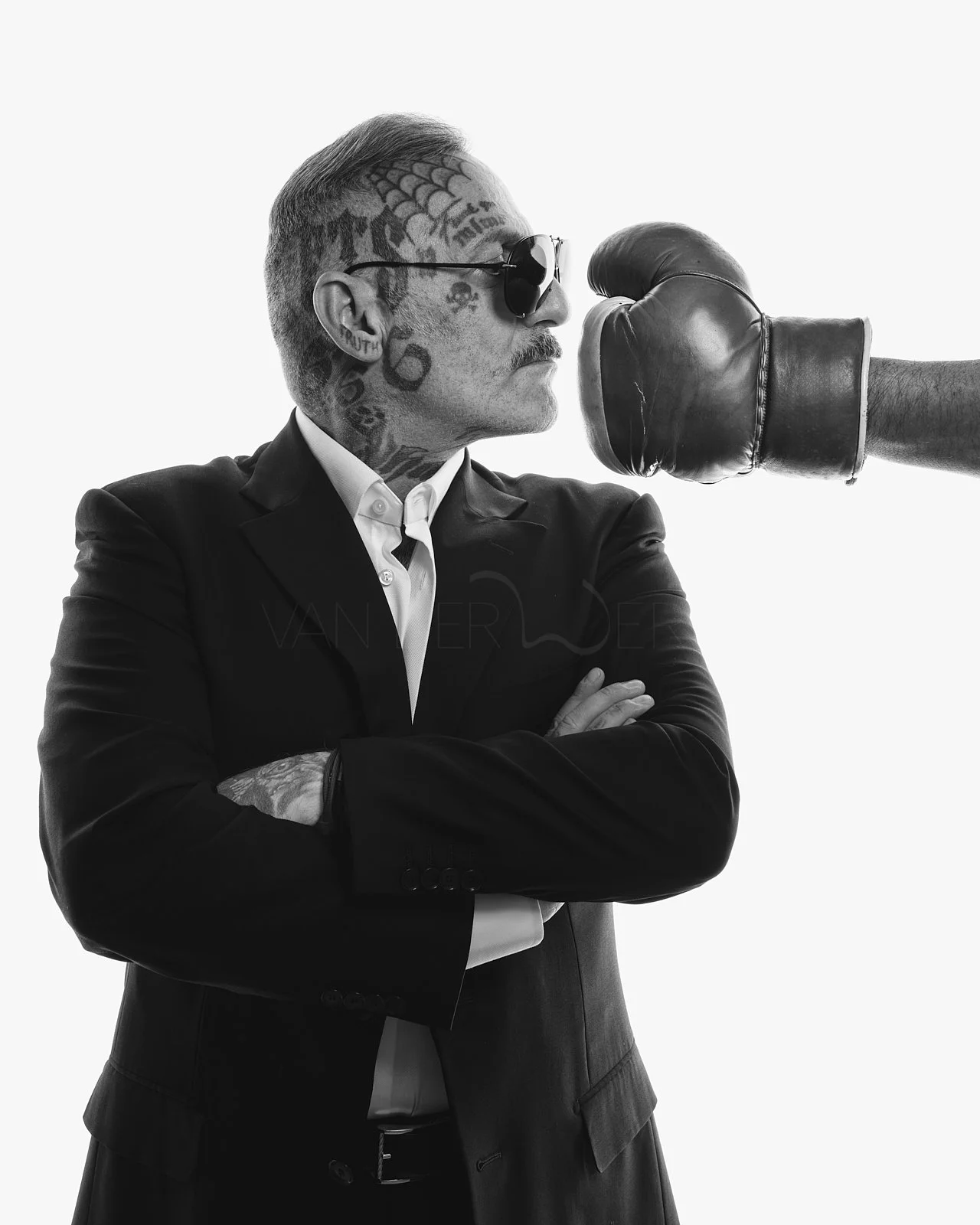 Black-and-white photo of a man with tattoos, wearing sunglasses and a suit, crossing his arms while a boxing glove is thrown at him.