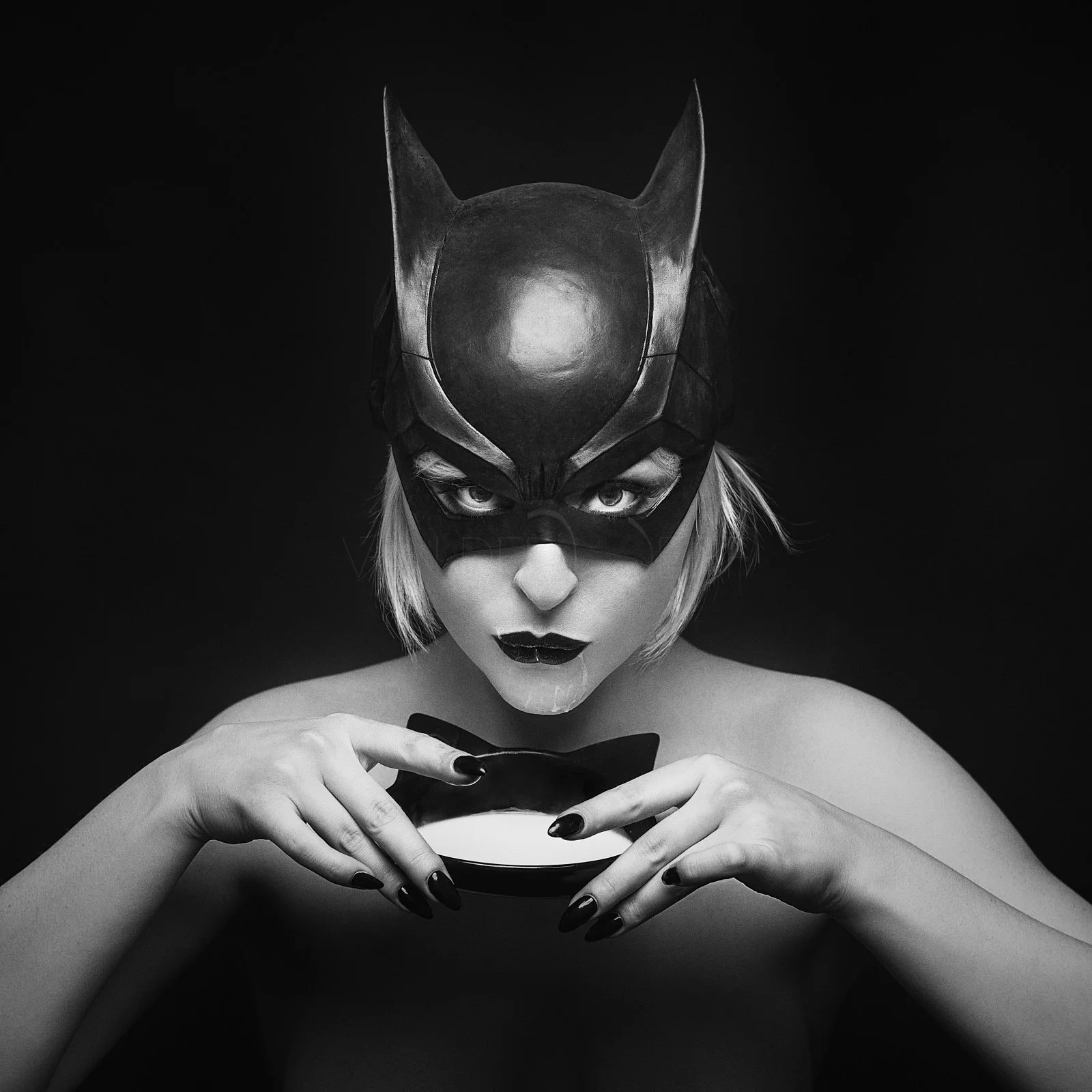 A person wearing a Batman mask with pointed ears and a black top, holding a small black bowl against a black background. The person has short hair, dark makeup, and black painted fingernails, and is staring directly at the camera.