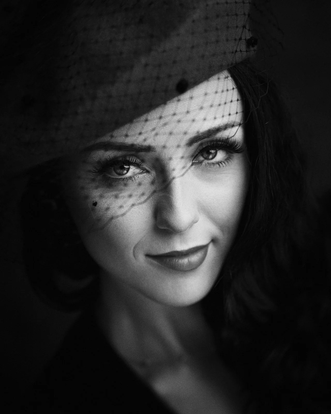 Black-and-white portrait of a woman wearing a wide-brimmed hat with netting, looking directly at the camera with a subtle smile