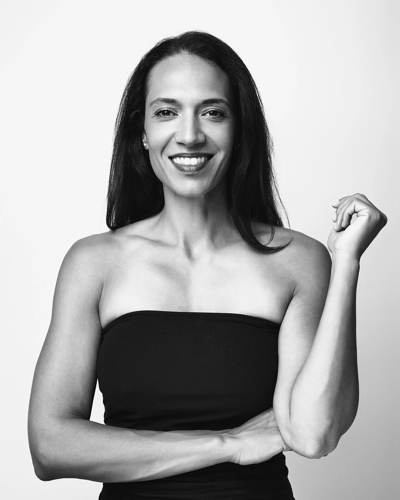 Black and white portrait of a smiling woman with long dark hair, wearing a strapless top, posing with one arm flexed.