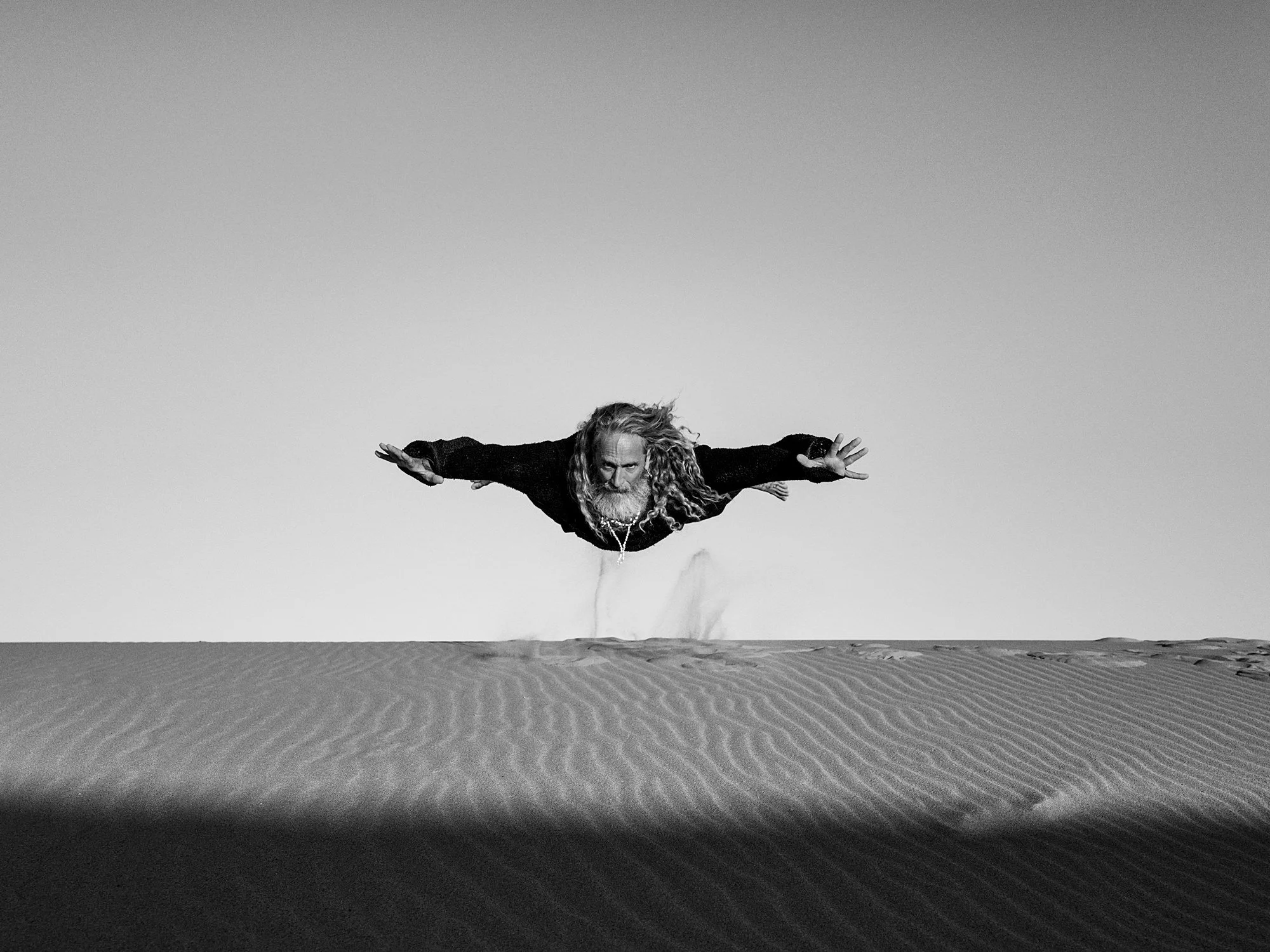 A man with long hair and a beard levitates above sand dunes in a desert, with arms extended horizontally and head facing downward, in a black-and-white photograph.