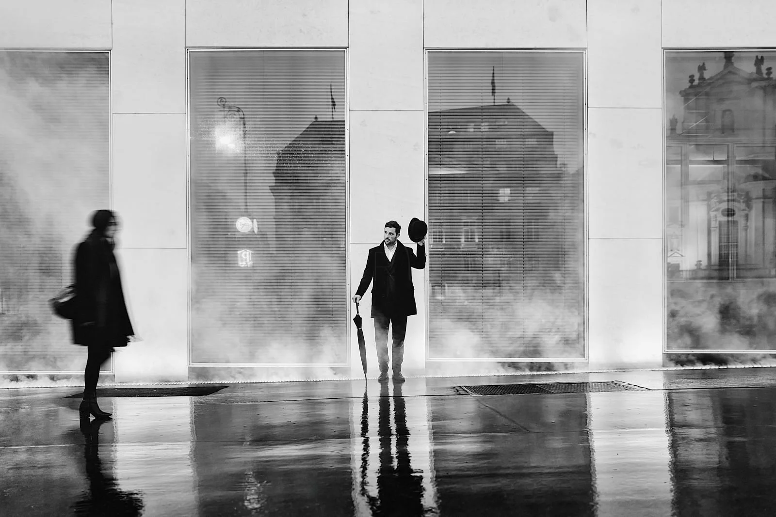 A man in a suit holding an umbrella and a hat stands in front of a large glass building with reflections, while a woman walks past in a coat, on a wet city street in black and white.
