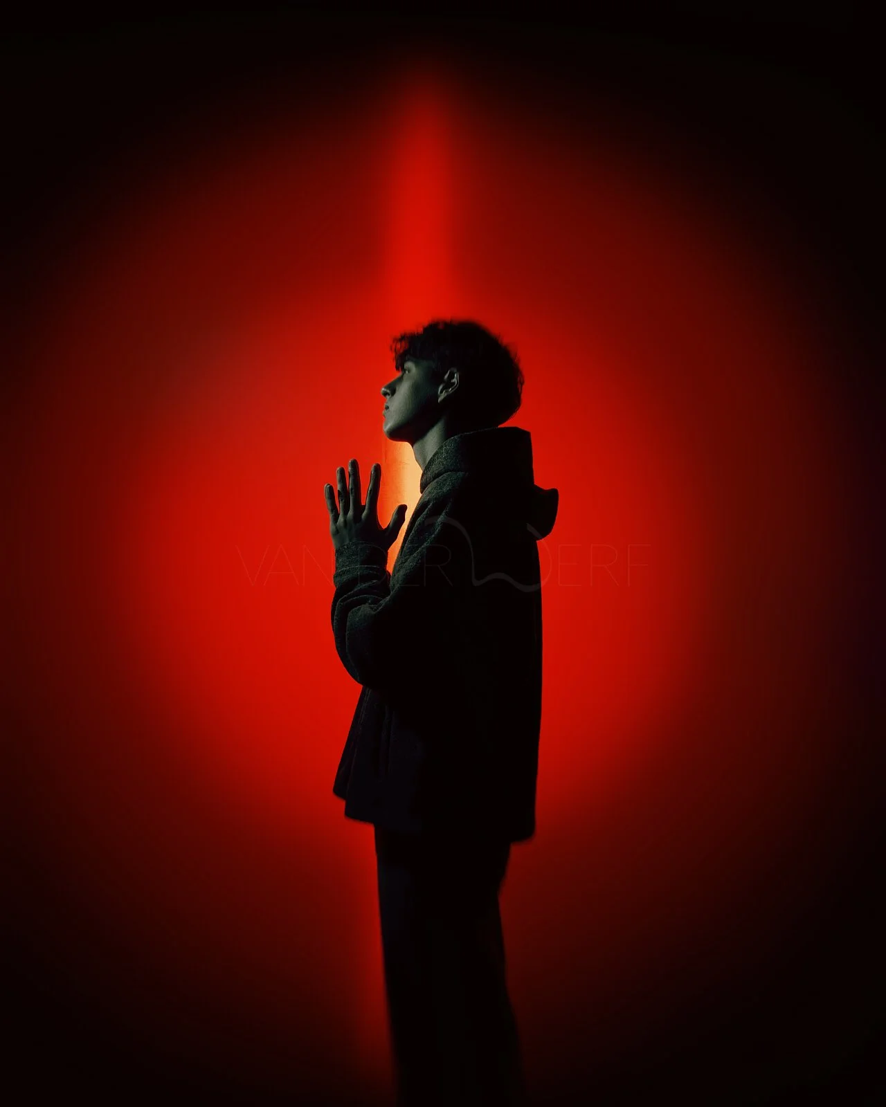 Silhouette of a young man with curly hair, standing in profile with hands pressed together in front of a red and black gradient background.