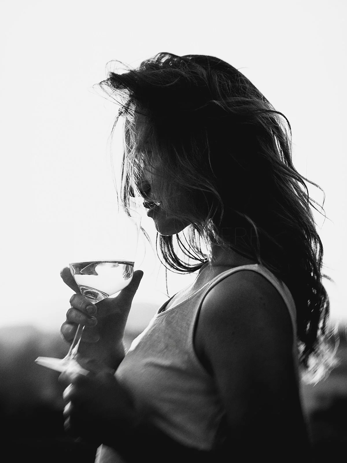 Silhouette of a woman with wavy hair holding a wine glass, looking down in profile, against a bright background.