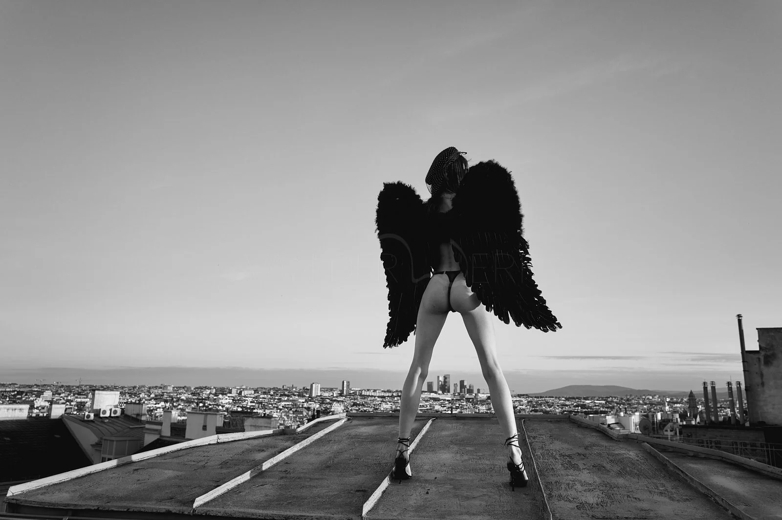 A person with large black angel wings, wearing high heels and minimal clothing, stands on a rooftop overlooking a cityscape in black and white.