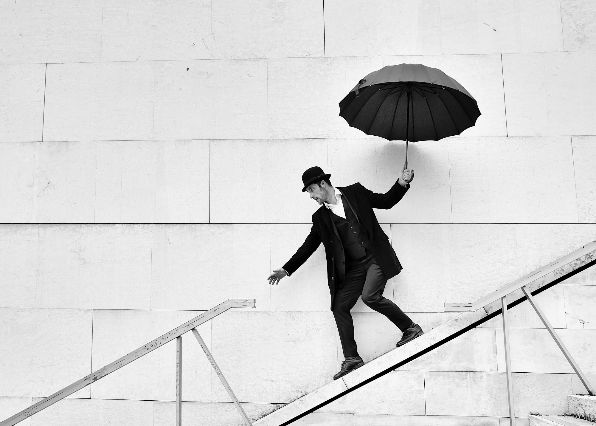 A man in a suit and bowler hat holding an umbrella appears to be falling or slipping down a staircase against a plain wall.