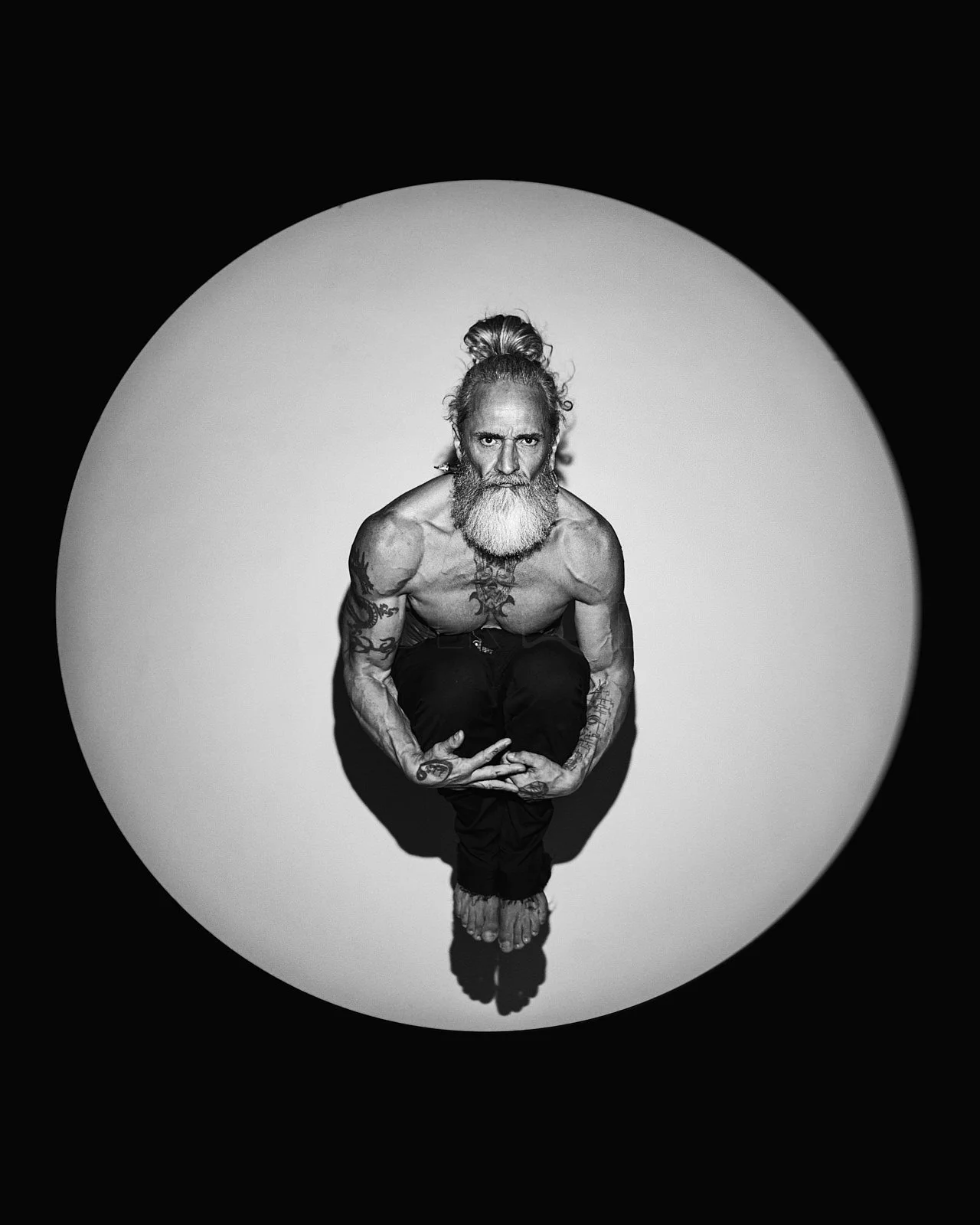 Black and white photo of a shirtless man with tattoos, a long beard, and long hair tied in a bun, squatting with their arms resting on their knees, looking directly at the camera, inside a circular spotlight against a dark background.