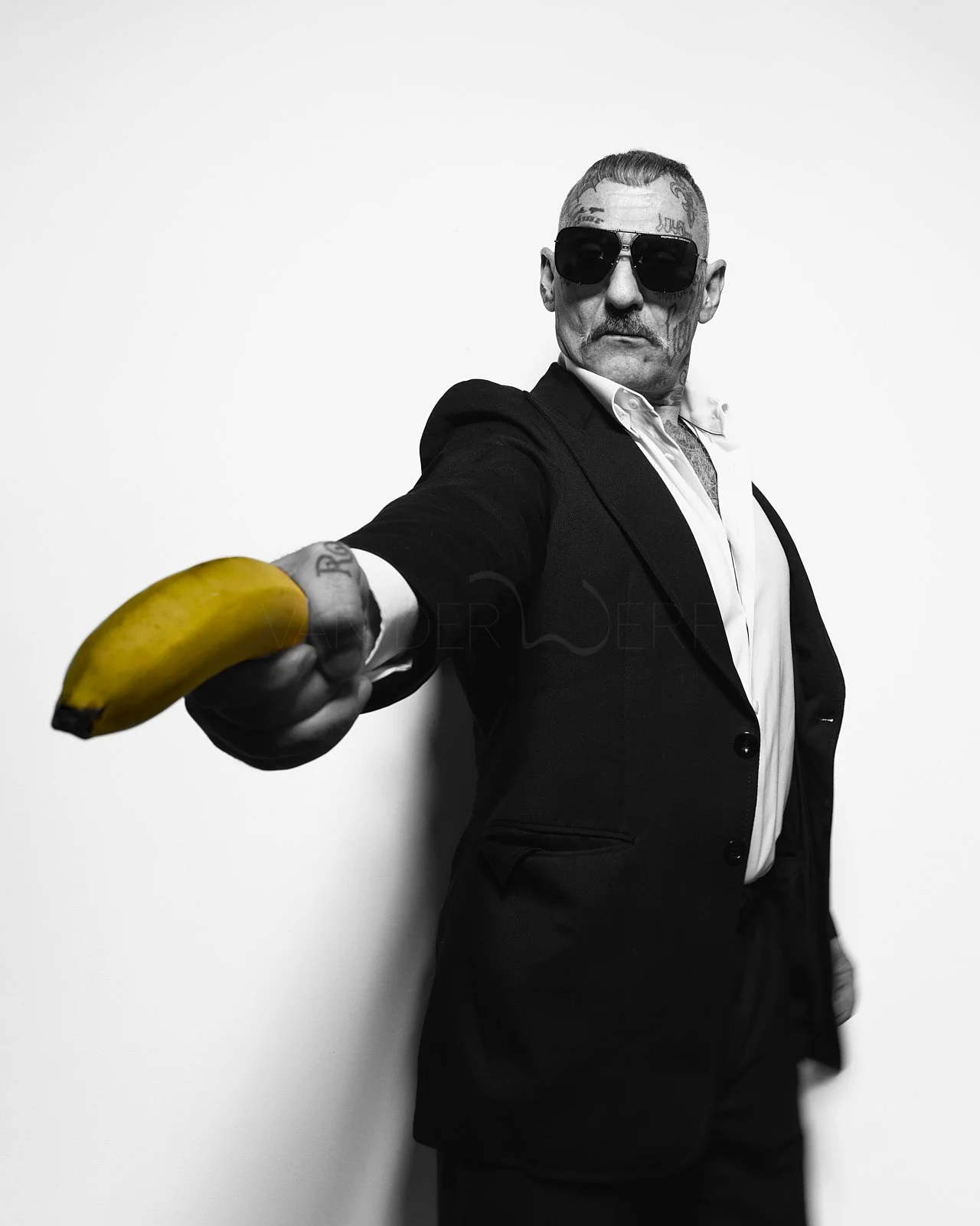 Black and white photo of a man in a suit and sunglasses holding a banana toward the camera.