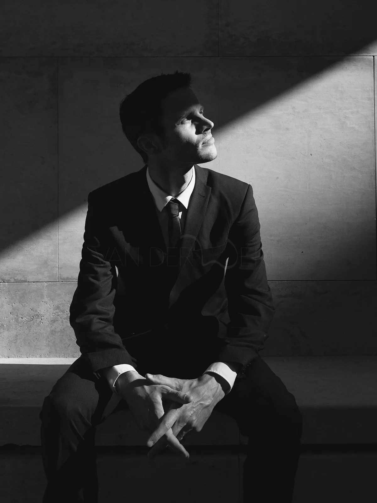 A man in a suit sitting on a bench with a dramatic light and shadow pattern on his face and background.