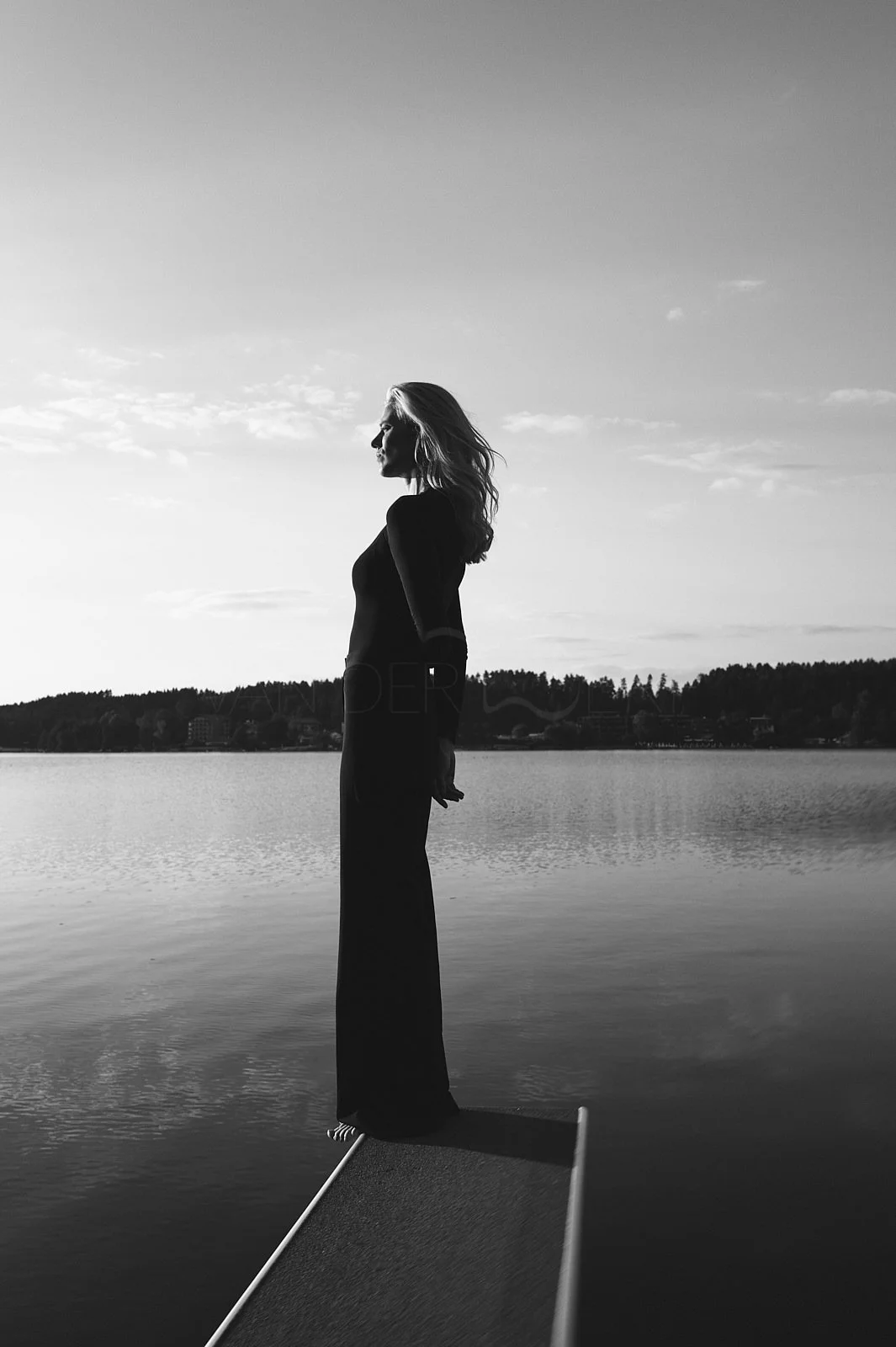 Silhouette of a woman standing on the edge of a dock by a lake, facing left, in black and white, with trees and buildings in the background.