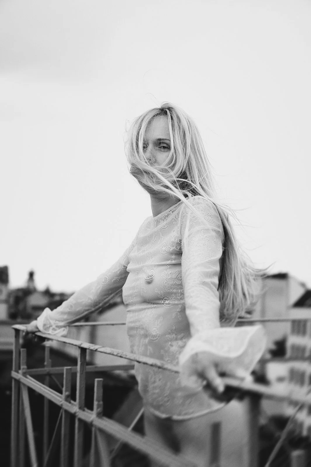 Black and white photo of a woman with long hair on a rooftop, holding onto a metal railing, with wind blowing her hair and wearing a lace long-sleeve top.