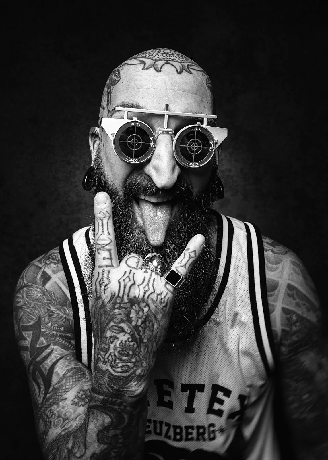 Man with tattoos, a beard, and glasses with target designs, sticking out his tongue and making a rock hand gesture.