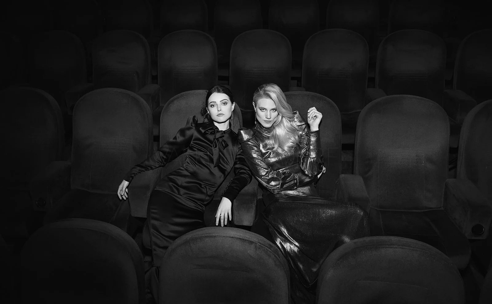Two women sitting in theater seats, one in a shiny black dress with a bow tie and the other in a metallic outfit, surrounded by empty theater seats, black and white photo.