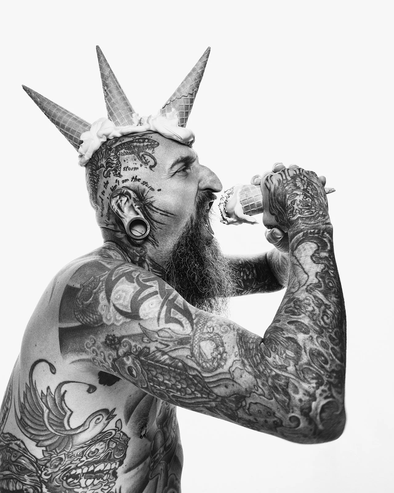 Tattooed man with a beard, wearing a cone hat resembling an ice cream cone, is licking a small ice cream cone. The image is in black and white.